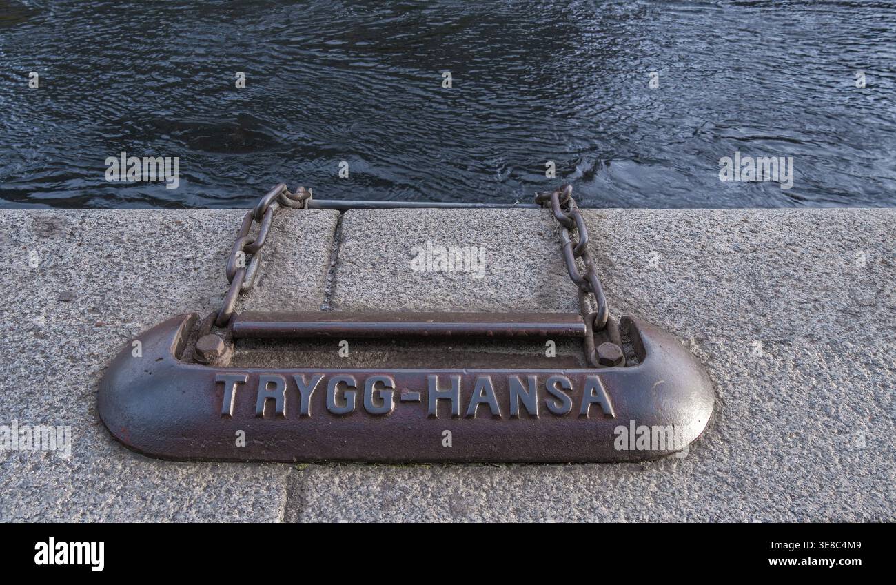 Stockholm, Suède - 11 octobre 2025 : borne d'amarrage avec chaîne sur un quai en béton à côté de l'eau ondulée, texte en relief “trygg-hansa”. DÉT. Quai. Maritime Banque D'Images