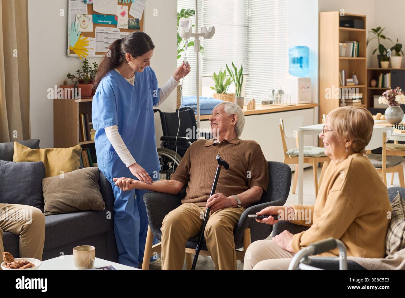 Homme caucasien senior recevant un traitement IV par une infirmière en maison de soins, assis avec une canne et interagissant avec le groupe. Utile pour les soins aux personnes âgées, les soins à domicile, le marketing de la vie assistée Banque D'Images