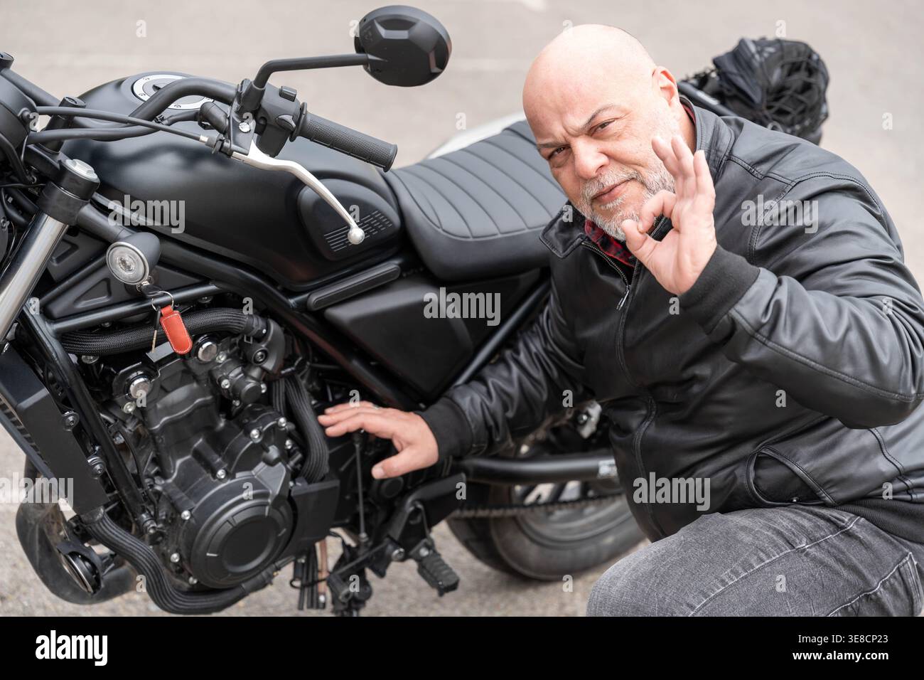 Homme chauve avec la barbe portant une veste en cuir noir, faisant un geste correct à côté de sa moto, montrant son approbation Banque D'Images