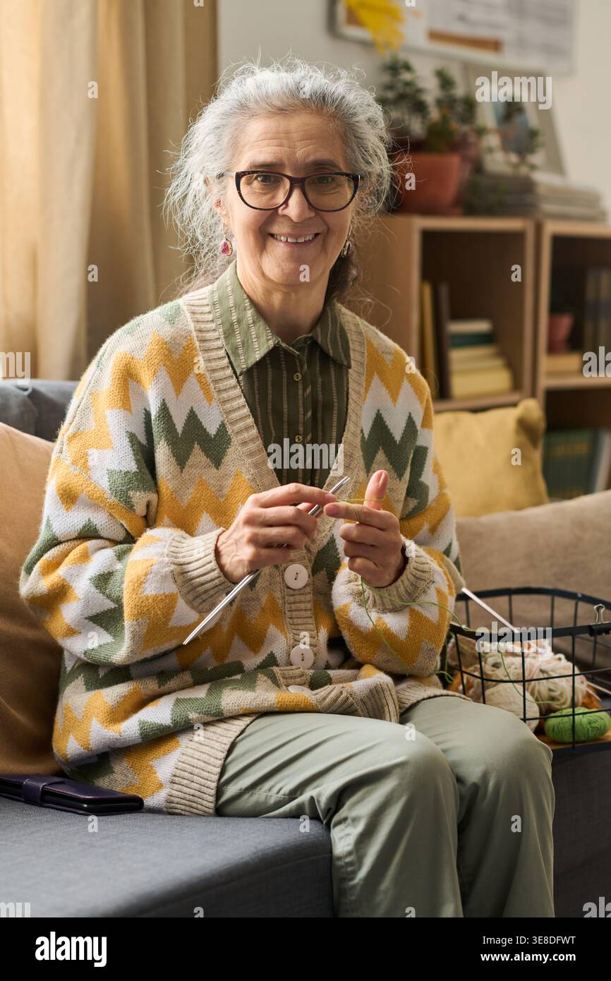 Portrait de femme senior caucasienne tricotant à la maison, souriant à la caméra et pratiquant un passe-temps créatif pour le concept de vieillissement actif. Utile pour le style de vie de retraite, le bien-être, le marketing des loisirs, vertical Banque D'Images