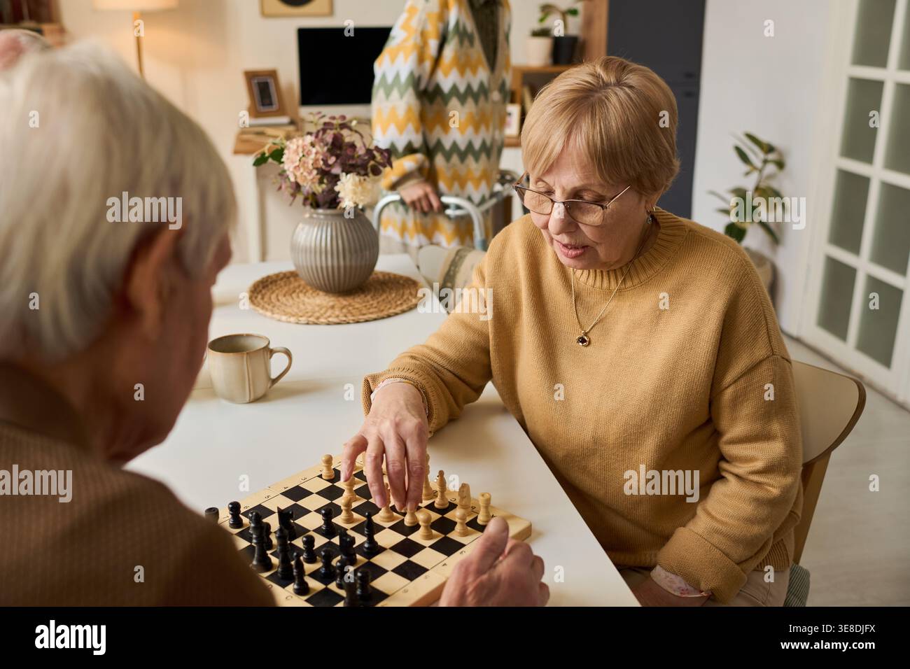 Femme caucasienne senior jouant aux échecs avec un homme âgé à la maison, soutenant l'activité cognitive et les soins sociaux. Utile pour le vieillissement, la prévention de la démence, le marketing de style de vie de retraite Banque D'Images