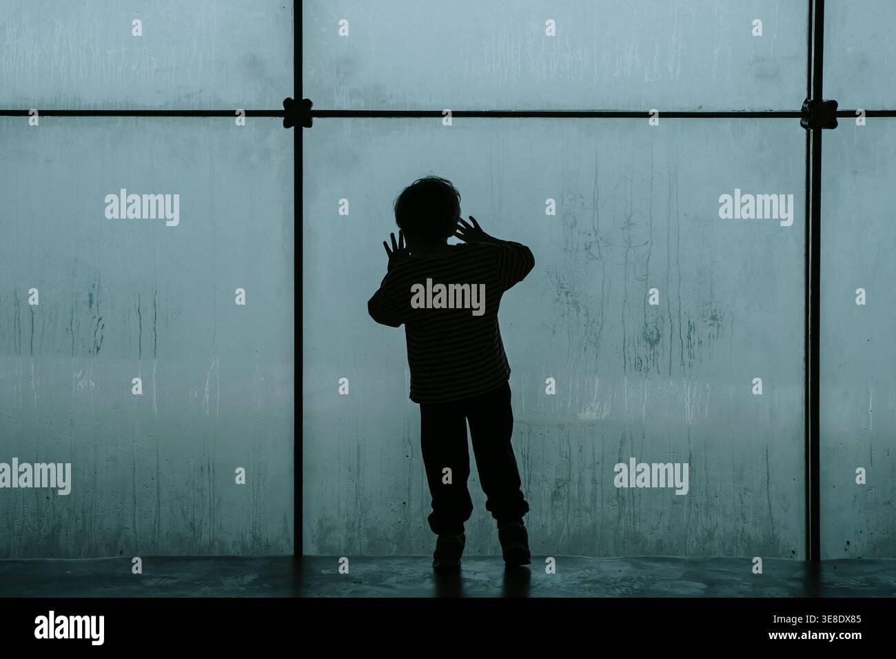 Silhouette d'un enfant solitaire regardant à travers une grande fenêtre embuée froide. Un travail de photographie conceptuelle explorant les thèmes de la dépression infantile, de la crise psychologique et du profond sentiment d’isolement et de tristesse. Photo de haute qualité Banque D'Images