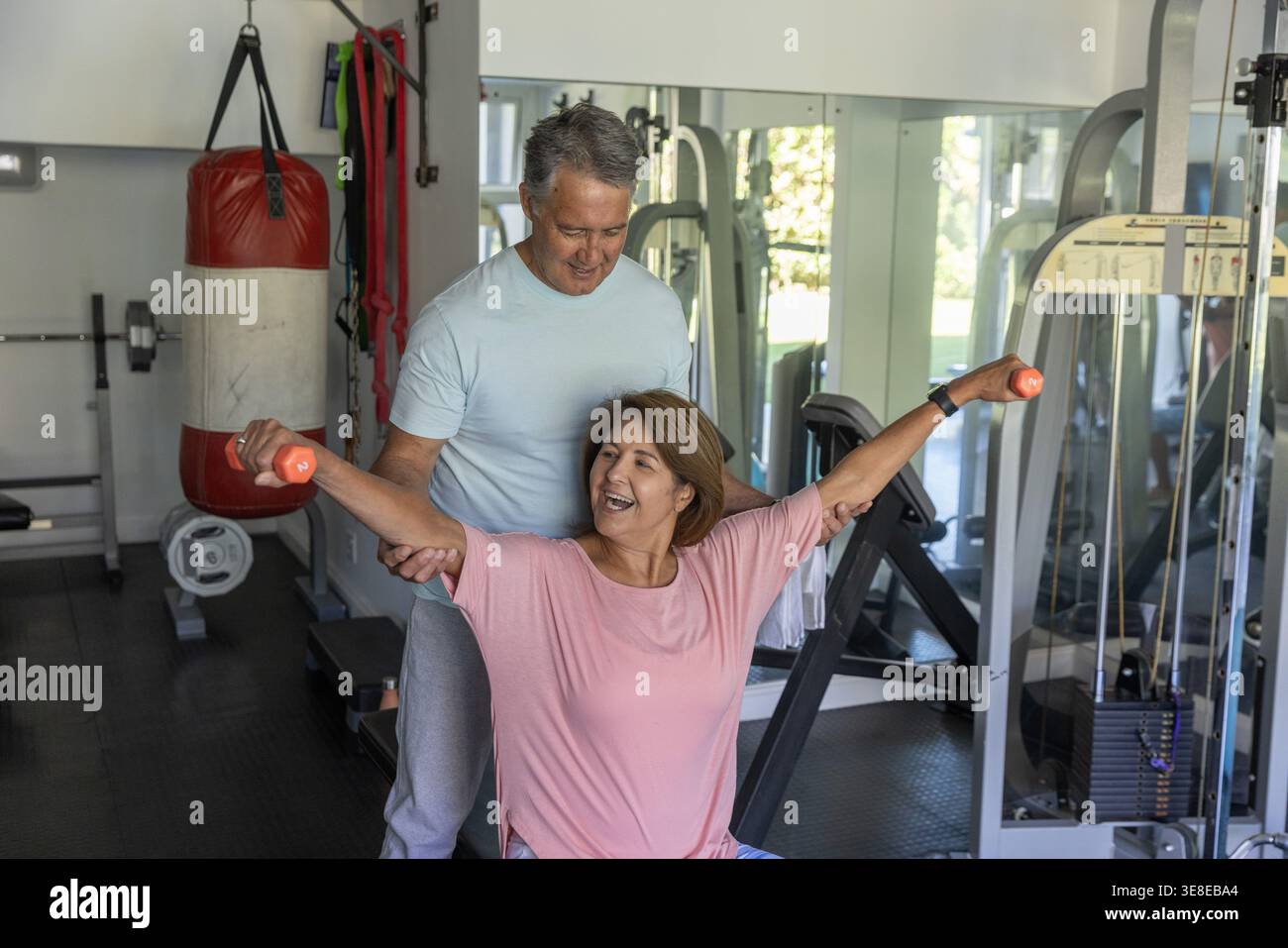 Femme senior en haut rose soulevant des haltères orange sur le banc tandis que l'homme soutenant les bras, les miroirs de gymnastique Banque D'Images