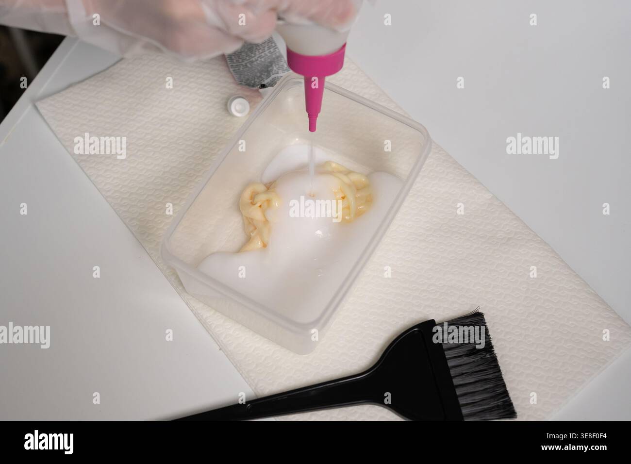la main gantée presse le traitement crémeux d'un tube dans un bol en plastique à côté d'une brosse et d'une bouteille. concept de coloration capillaire de préparation de salon de beauté Banque D'Images