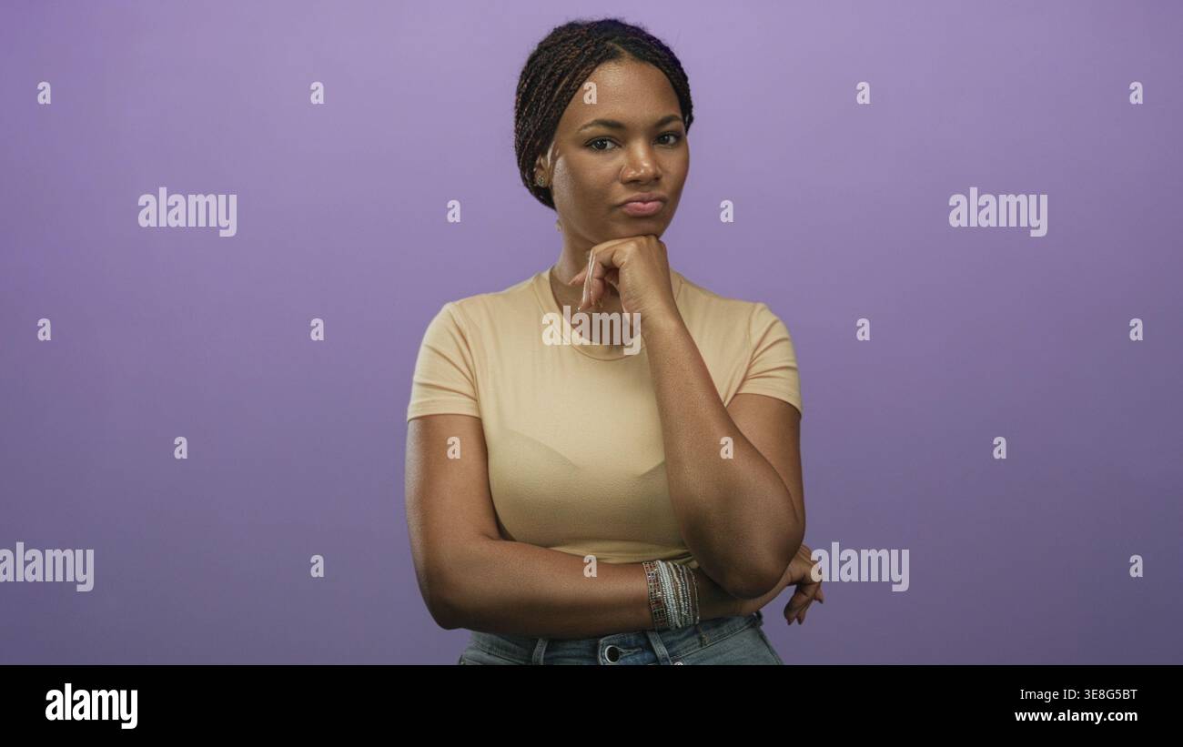 Femme avec la main sur le menton et les bras pliés debout dans le studio à côté du mur violet portant un tee-shirt beige et un Jean denim ; pensée contemplative. Banque D'Images