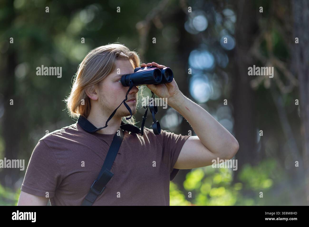 Plan de profil latéral d'un jeune homme aux longs cheveux blonds regardant à travers une paire de jumelles dans un cadre forestier. Banque D'Images