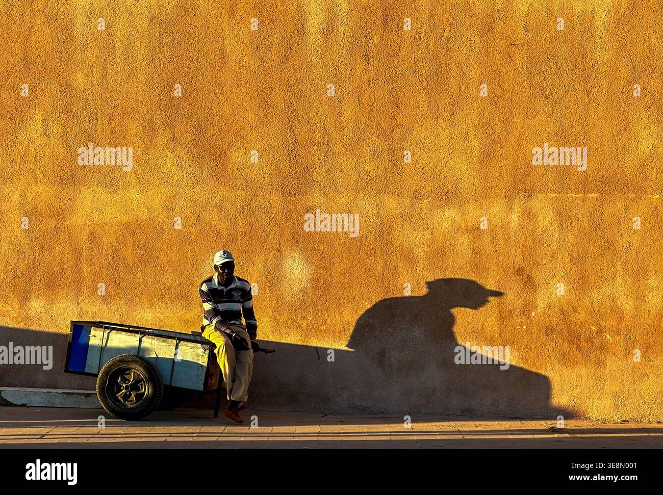Vendeur de rue se reposant à côté d'un chariot jetant une longue ombre sur un mur ensoleillé à Essaouira, Maroc Banque D'Images