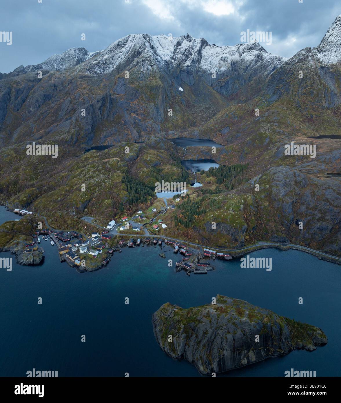 Vue aérienne du village de pêcheurs de Nusfjord avec des cabanes rouges traditionnelles, entouré de montagnes escarpées enneigées et des eaux sombres du fjord Nusfjord, Nordla Banque D'Images