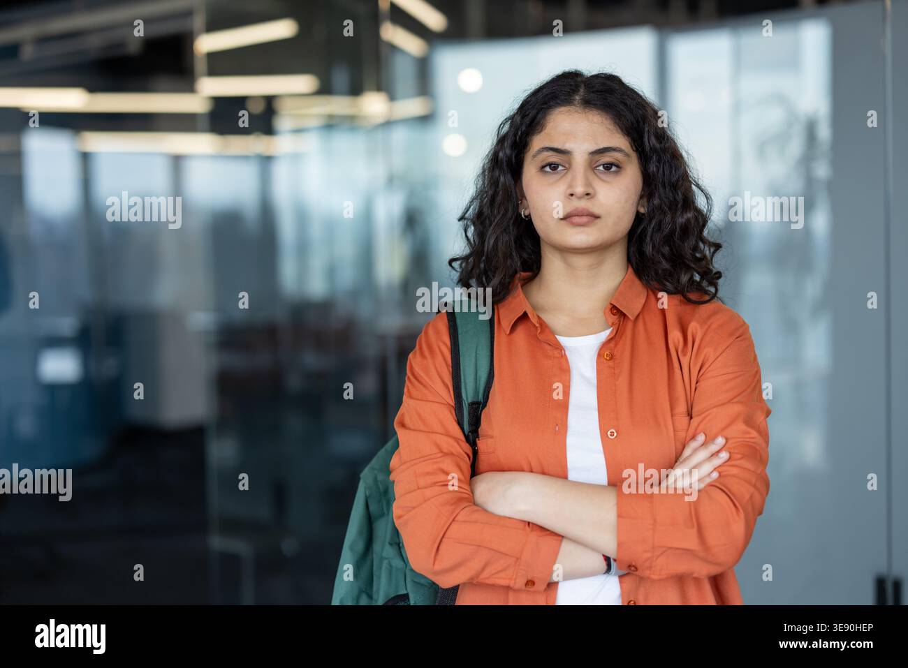 Jeune femme debout avec une expression sérieuse, portant une chemise orange et un sac à dos, véhiculant détermination et ambition dans un universitaire ou une profession Banque D'Images