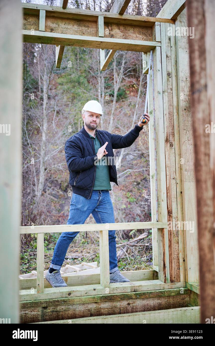Travailleur de la construction mesurant la hauteur du mur avec du ruban à l'intérieur de la maison à ossature en bois, montrant le geste OK tout en confirmant les dimensions correctes pendant le processus de construction Banque D'Images