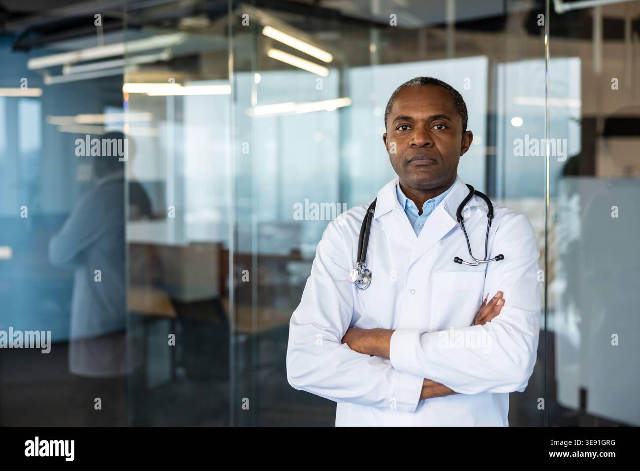 Médecin masculin expérimenté debout avec confiance avec les bras croisés, portant une blouse de laboratoire blanche et un stéthoscope, représentant le professionnalisme de la santé et l'expertise médicale dans un cadre de bureau Banque D'Images