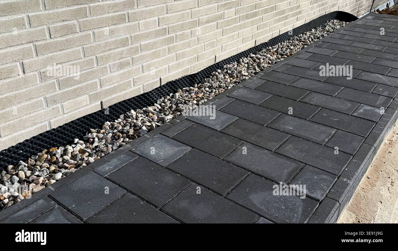 Passerelle pavée faite de blocs de béton gris foncé le long d'un mur de briques texturées avec du gravier et du drainage visible dans l'aménagement paysager Banque D'Images