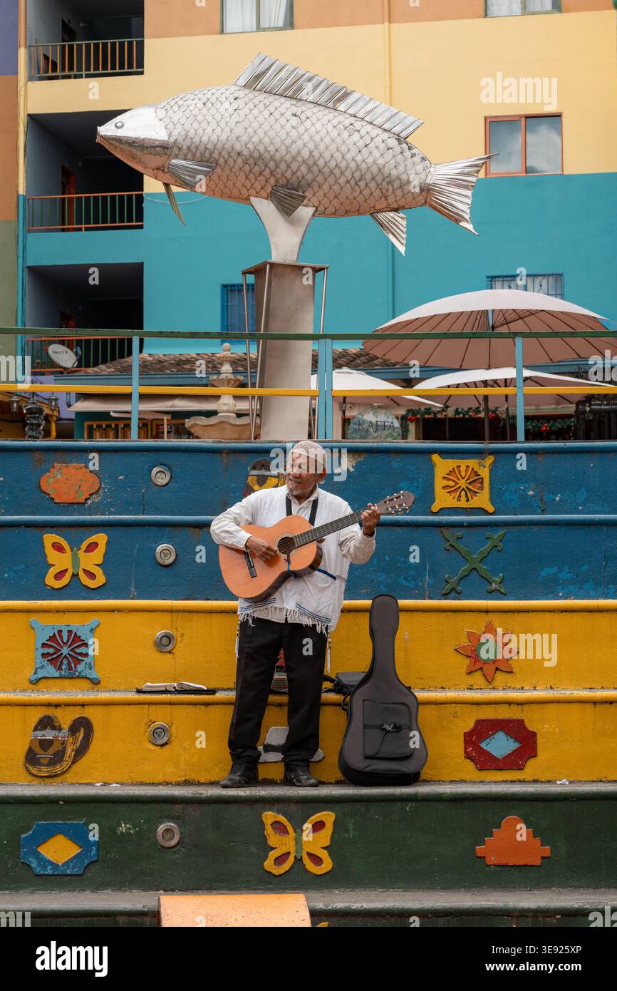 Musicien de rue jouant de la guitare pour une foule dans une place animée, avec une grande sculpture de poisson, zócalos coloré et architecte colombien traditionnel Banque D'Images