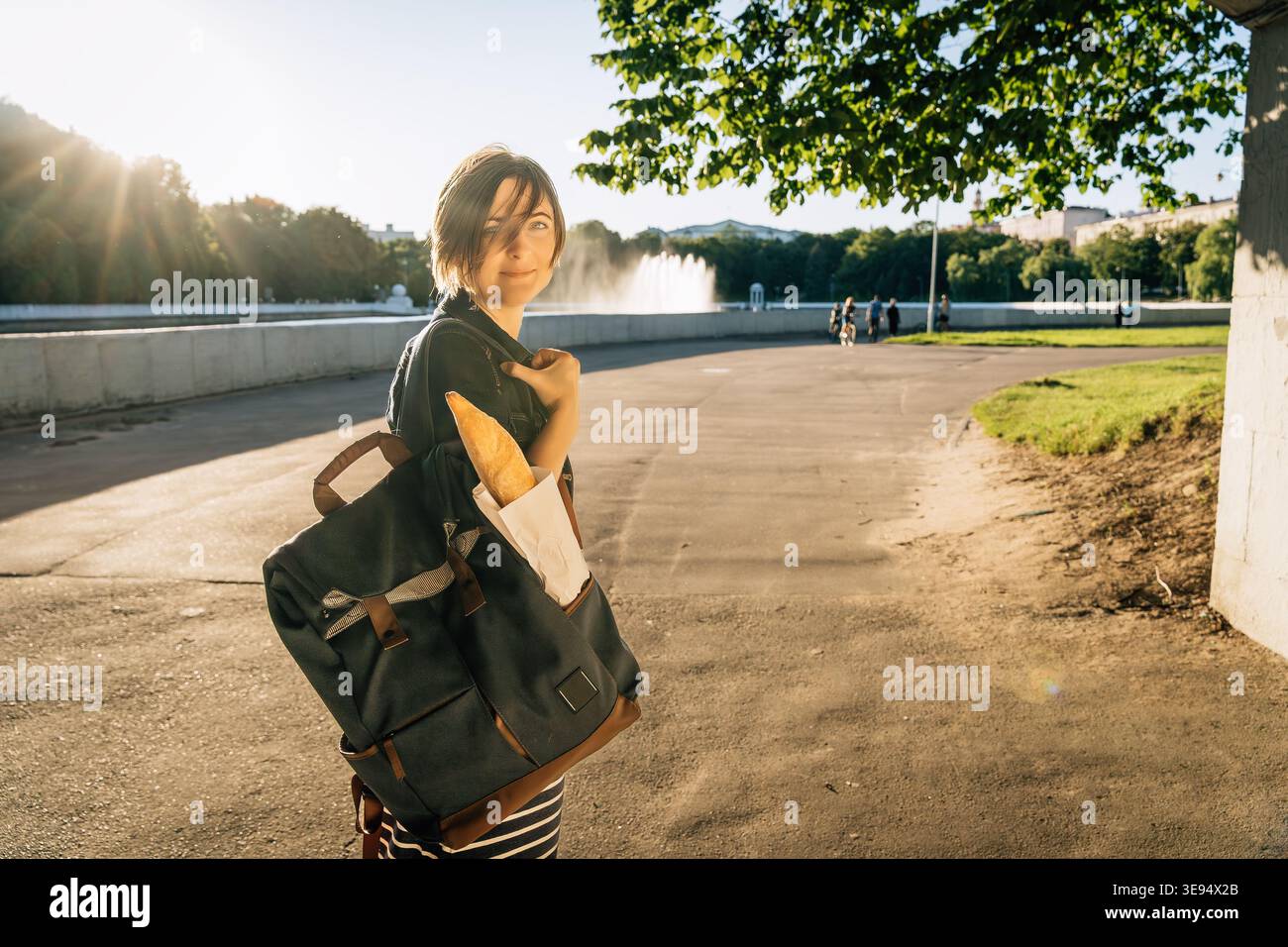 Femme souriante 40 avec sac à dos portant baguette dans le parc Banque D'Images