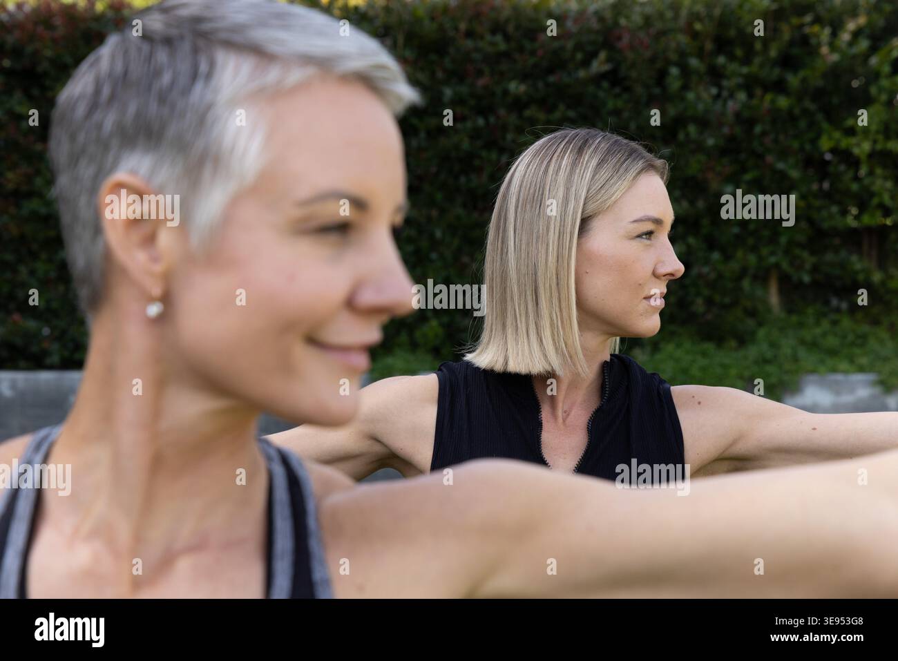Amies féminines s'étirant sur la pelouse herbeuse dans la cour arrière portant des débardeurs près de la haie taillée et du mur. Extérieur, fitness, yoga, stretch, duo, nature, bien-être Banque D'Images