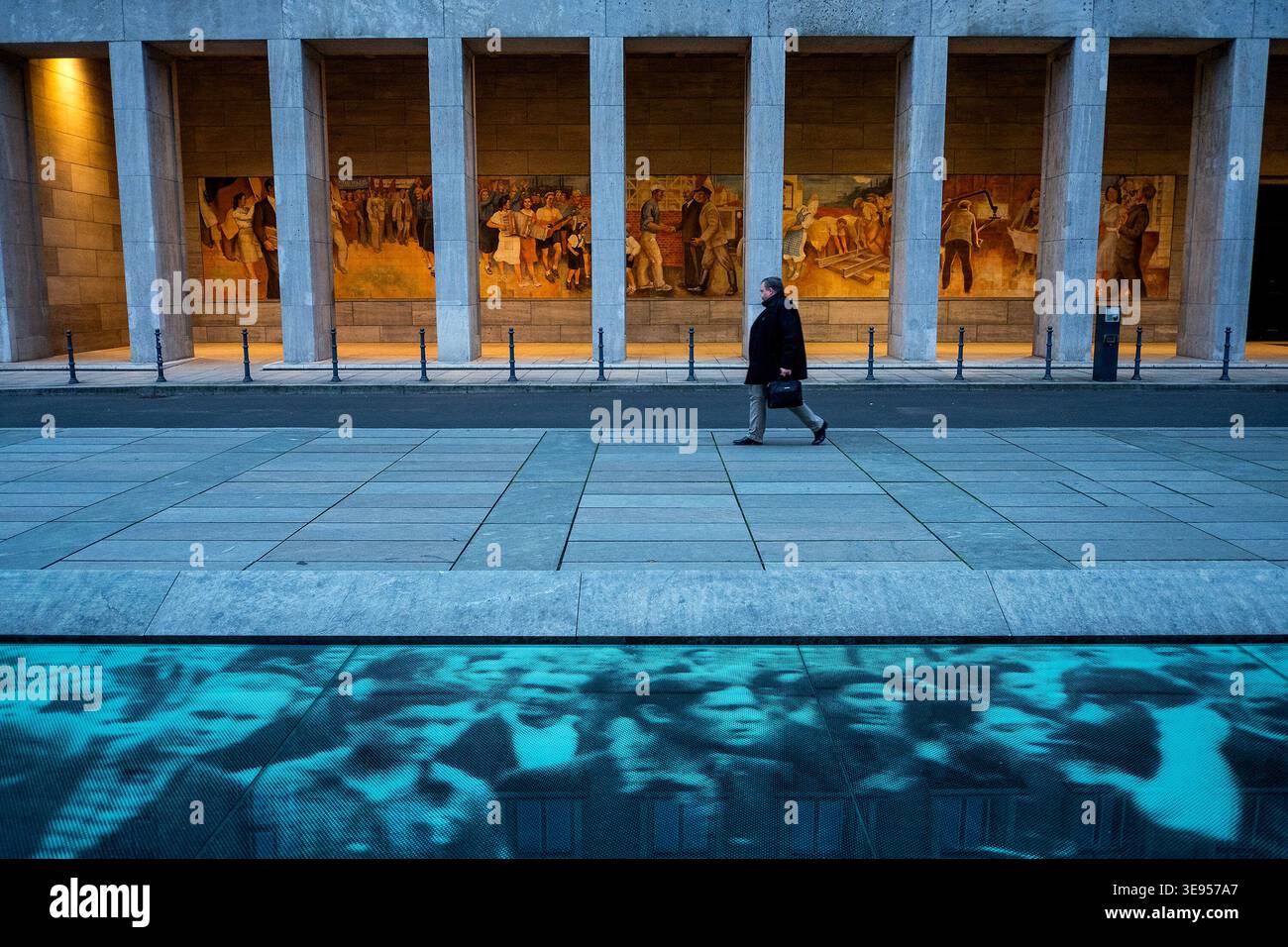 Une personne traverse une place devant un bâtiment avec des colonnes et des peintures murales. Banque D'Images