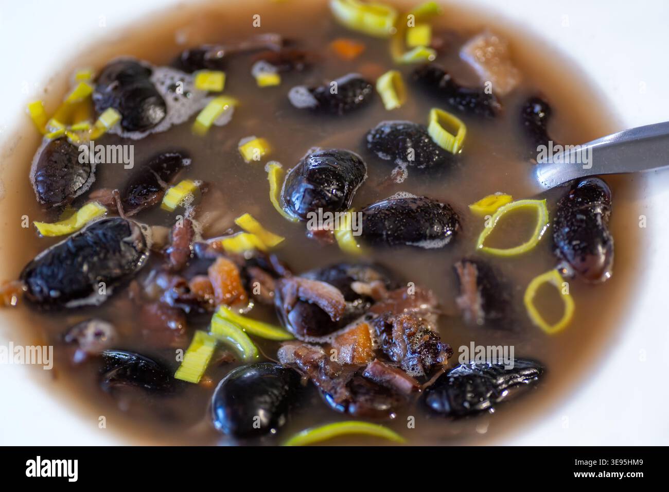 Soupe de haricots noirs avec légumes et poireaux tranchés dans une assiette blanche, cuillère, gros plan. Banque D'Images