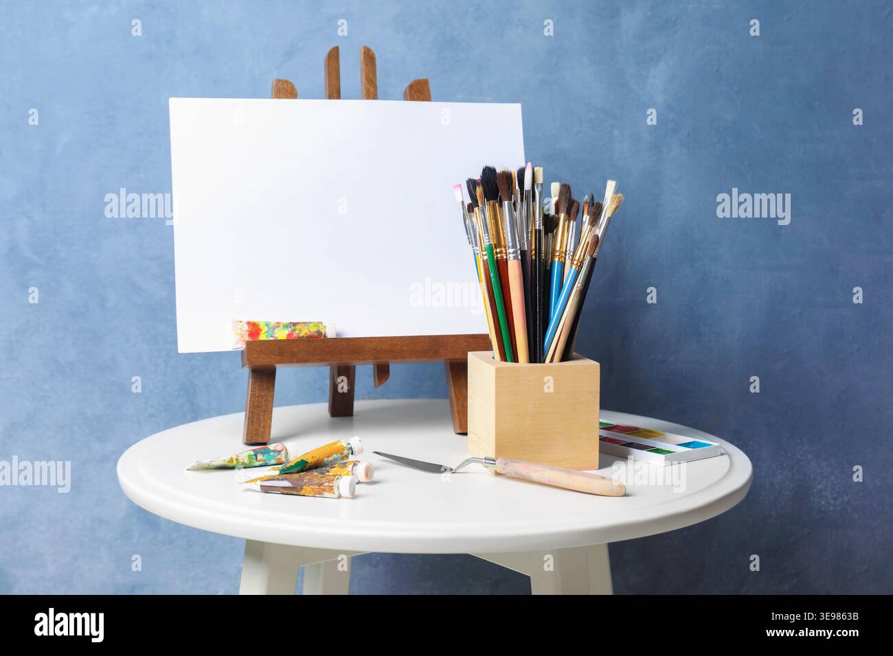 Pinceaux, couteau, peintures et chevalet avec toile sur table près du mur bleu Banque D'Images