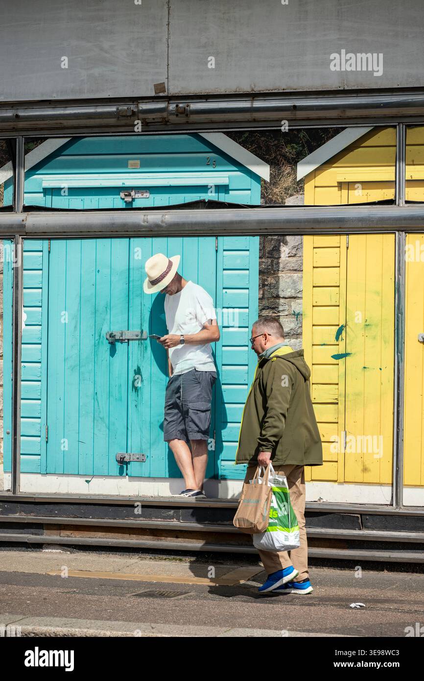 Homme plus âgé transportant des achats marchant devant un grand téléphone mobile publicitaire amassant avec un homme plus jeune utilisant un smartphone. Banque D'Images