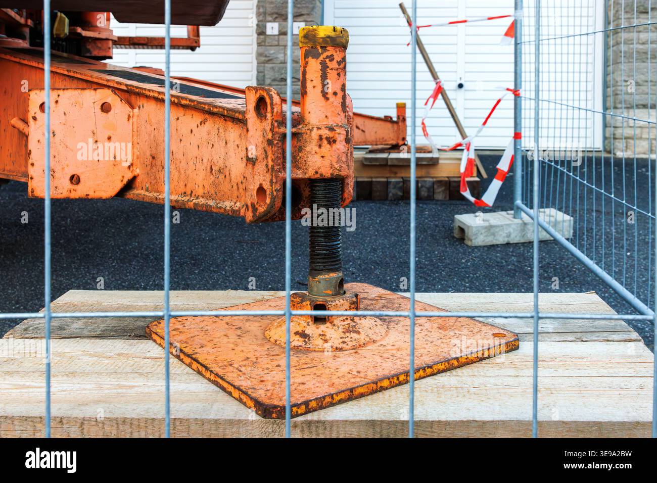 Gros plan d'un pied de support de stabilisateur de grue rouillé sur une plaque de base métallique à travers une clôture de sécurité temporaire sur un chantier routier. Banque D'Images