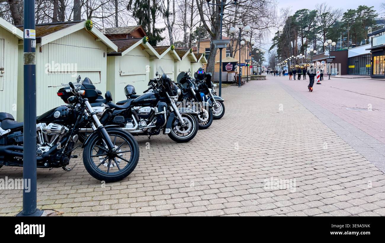 Riga, Lettonie - 29 mars 2026 : des motos alignées sur un chemin pavé à côté de cabines en bois, avec des piétons se promenant dans le fond d'une série Banque D'Images
