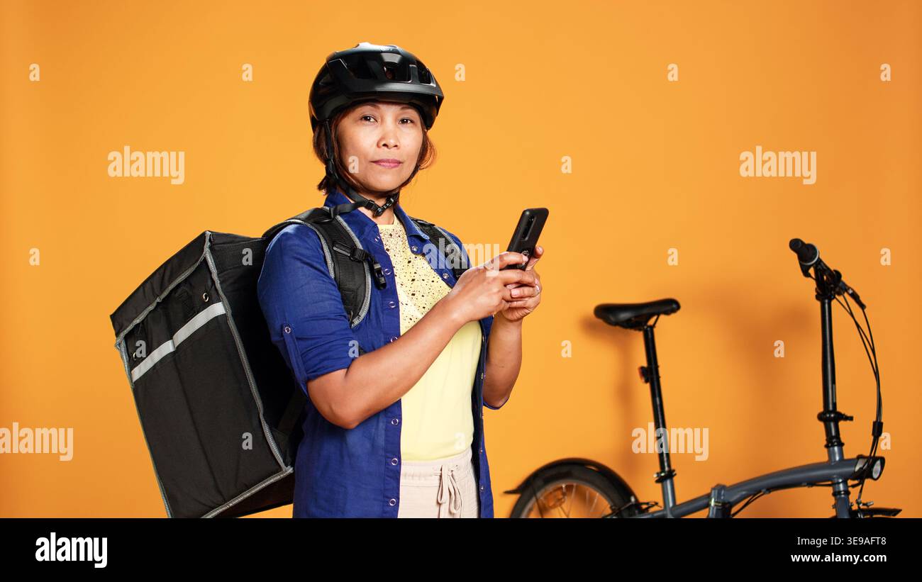 Portrait d'un coursier en attente, à la recherche dans l'application téléphonique du prochain emplacement de commande client. Femme BIPOC livrant de la nourriture avec bycicle en utilisant le sac thermique, isolé sur fond de studio Banque D'Images