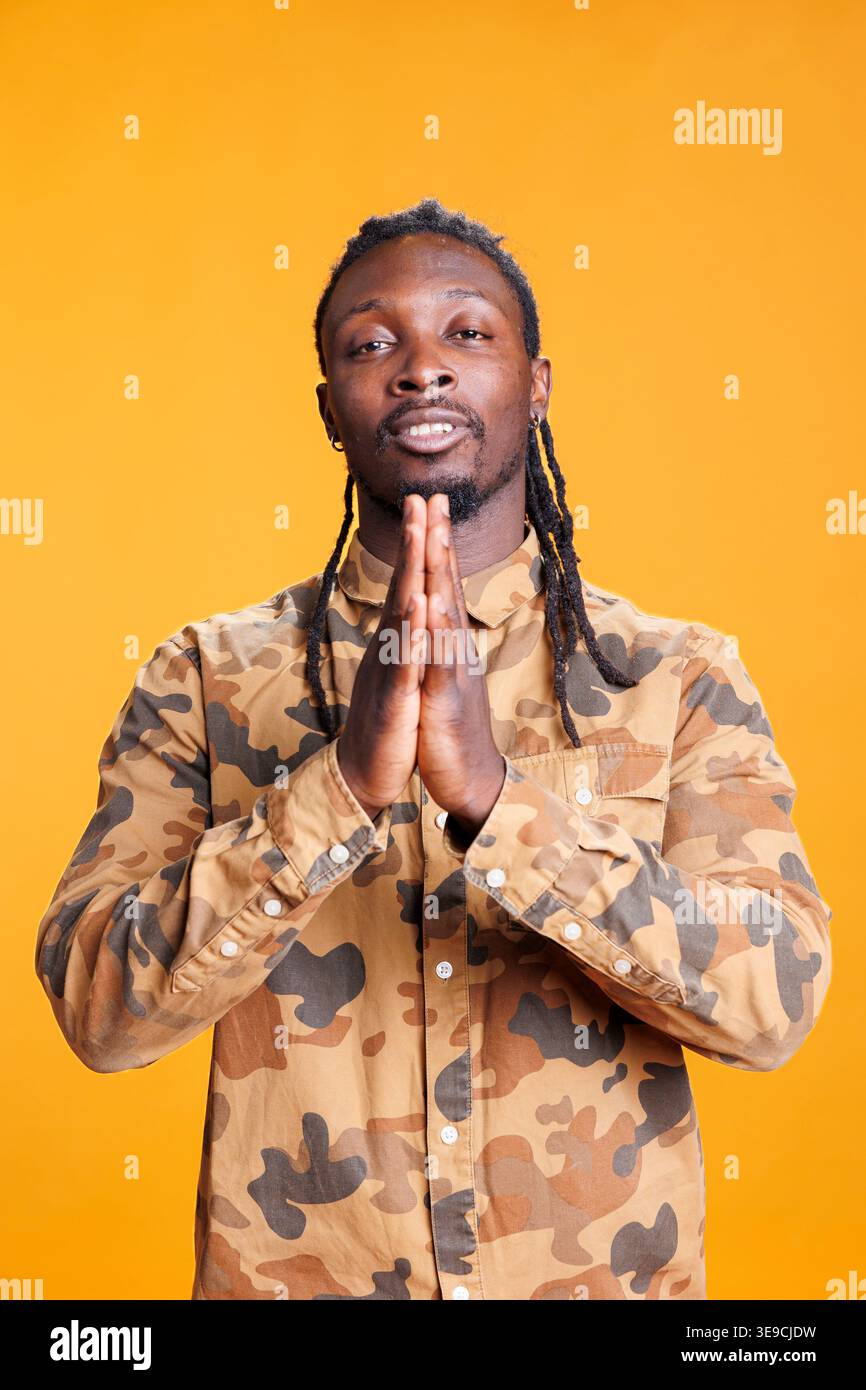 Portrait d'un homme paisible priant dieu demandant pardon tout en se tenant dans un studio avec fond jaune. Personne religieuse afro-américaine tenant les paumes ensemble dans Pray. Concept de religion Banque D'Images
