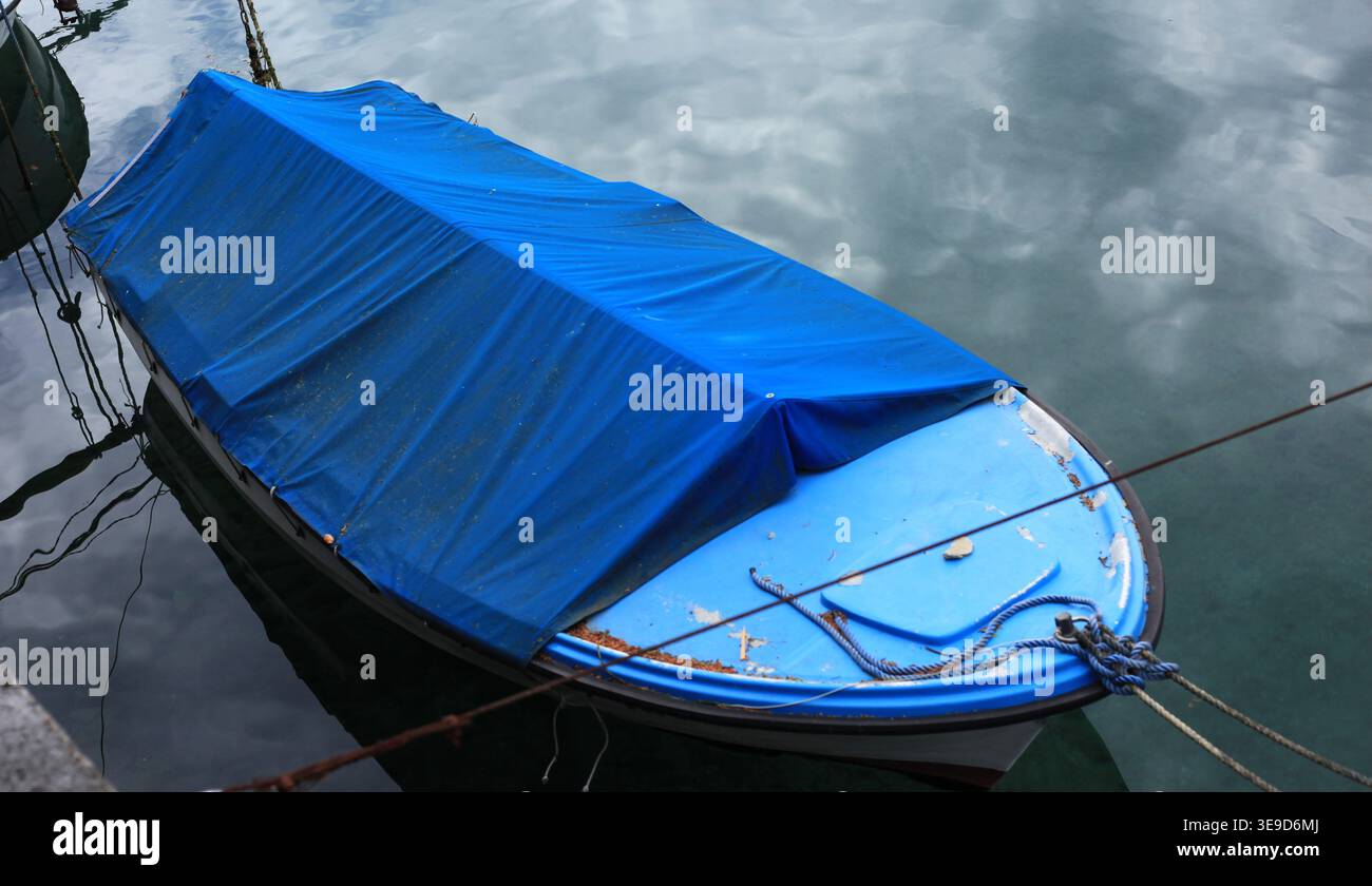 Un seul bateau de pêche bleu avec une couverture en toile amarré dans un port calme, reflétant le ciel. Banque D'Images