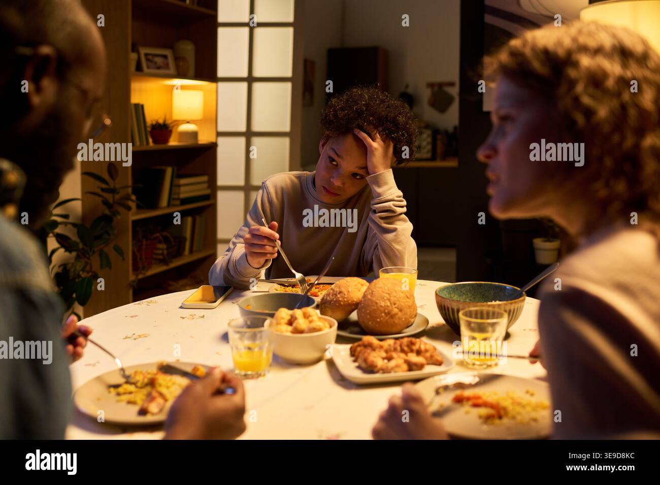 Adolescent assis à table, mangeant à contrecœur alors que les parents font face à une conversation familiale tendue à la maison. Convient pour le rôle parental, les conflits familiaux, le stress des adolescents, les campagnes de conseil Banque D'Images