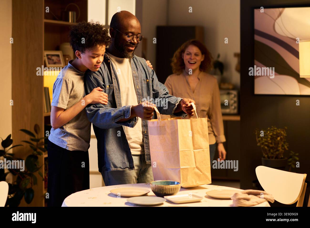 Père afro-américain et fils adolescent déballant les sacs à emporter à la table à manger tandis que la mère caucasienne approche, montrant la routine des repas de famille. Idéal pour la livraison de nourriture Banque D'Images