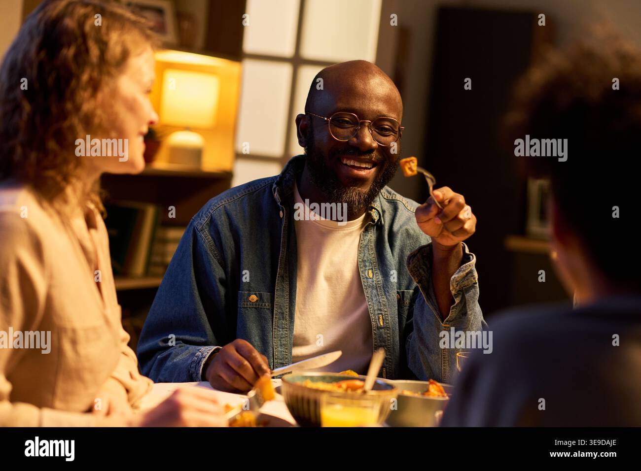Jeune homme adulte noir partageant le repas avec des amis à la table à manger, souriant et mangeant pendant la réunion sociale. Utile pour l'amitié, la communauté, la restauration à domicile, le marketing de style de vie Banque D'Images