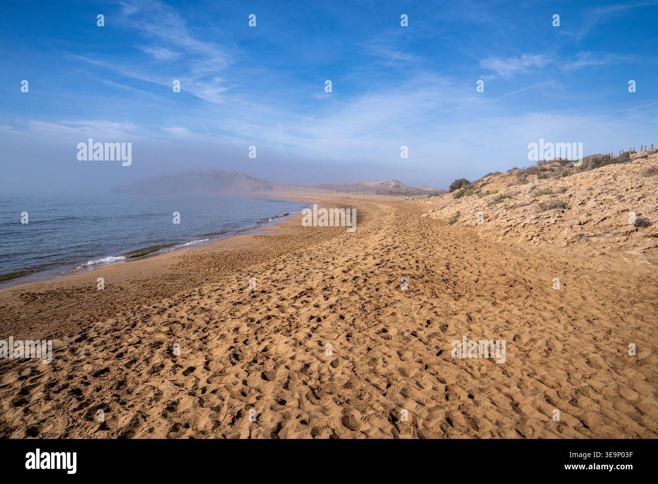 Vue panoramique sur la plage immaculée de Calblanque en Espagne. Le sable doré est marqué par des empreintes de pas et flanqué de dunes fossiles, tandis qu'un promontoire s'efface Banque D'Images