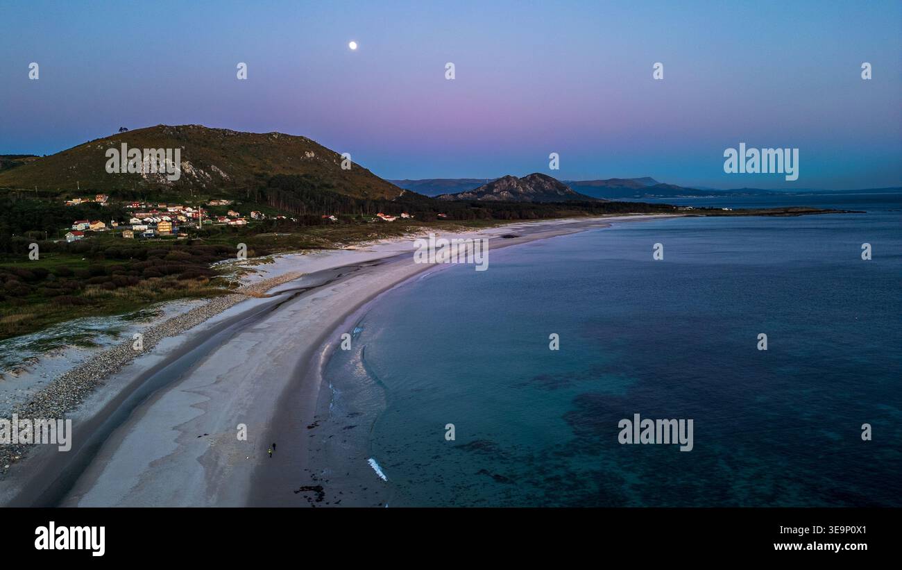 Une vue aérienne spectaculaire capture la plage de Lariño sous la lumière de la pleine lune. Le ciel crépusculaire violet pend au-dessus des montagnes et du village, tandis que th Banque D'Images