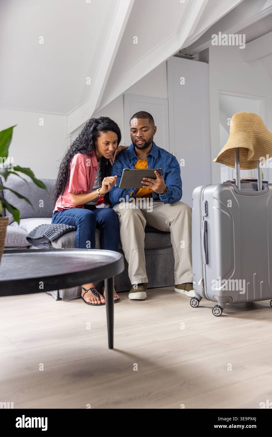 Couple diversifié assis sur le canapé gris dans le loft, étudiant tablette planifier voyage près de la valise avec chapeau Banque D'Images
