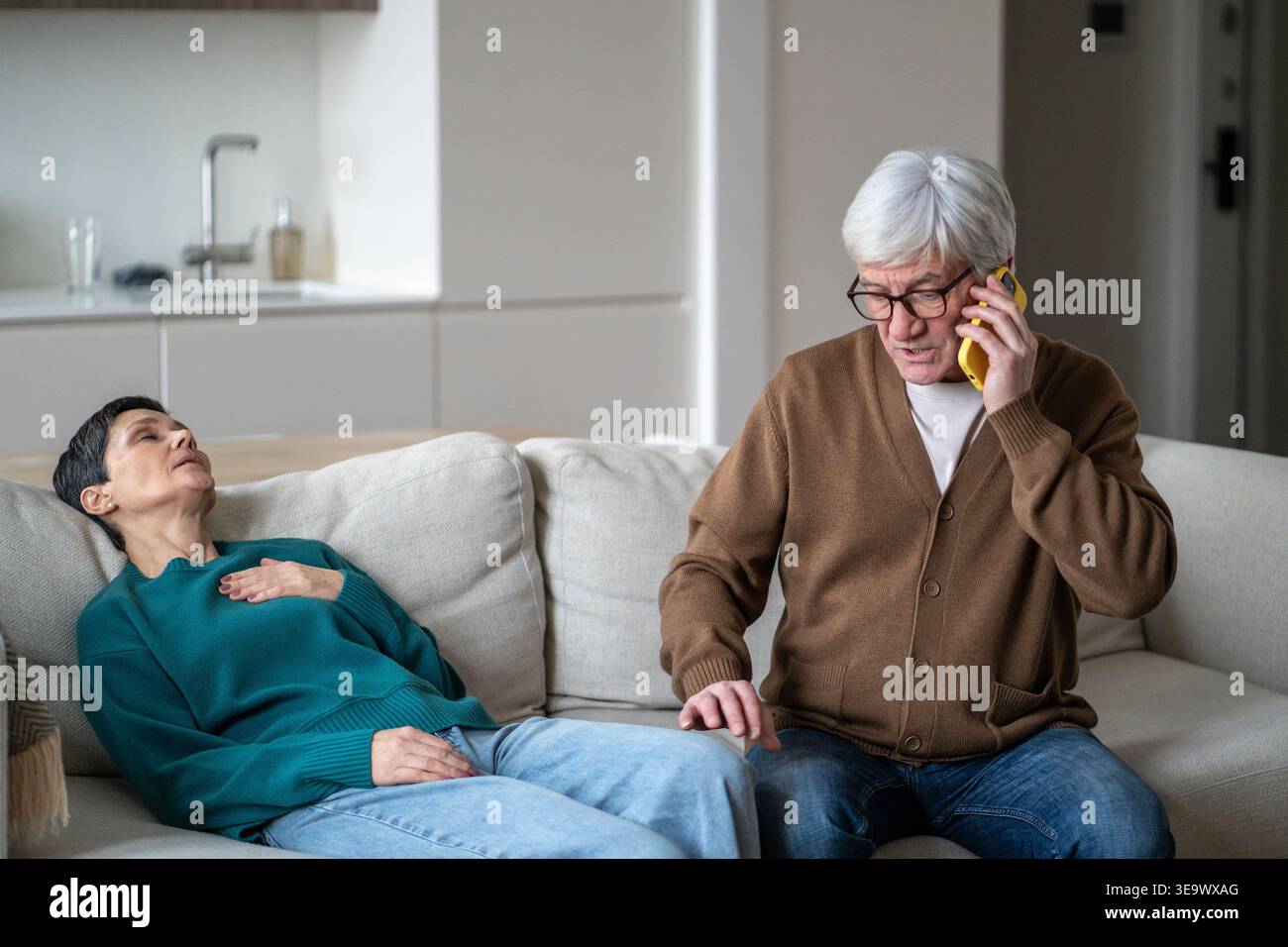 Femme âgée couchée sur le canapé dans la chambre avec douleur dans la poitrine tandis que le mari parle au médecin au téléphone. Banque D'Images