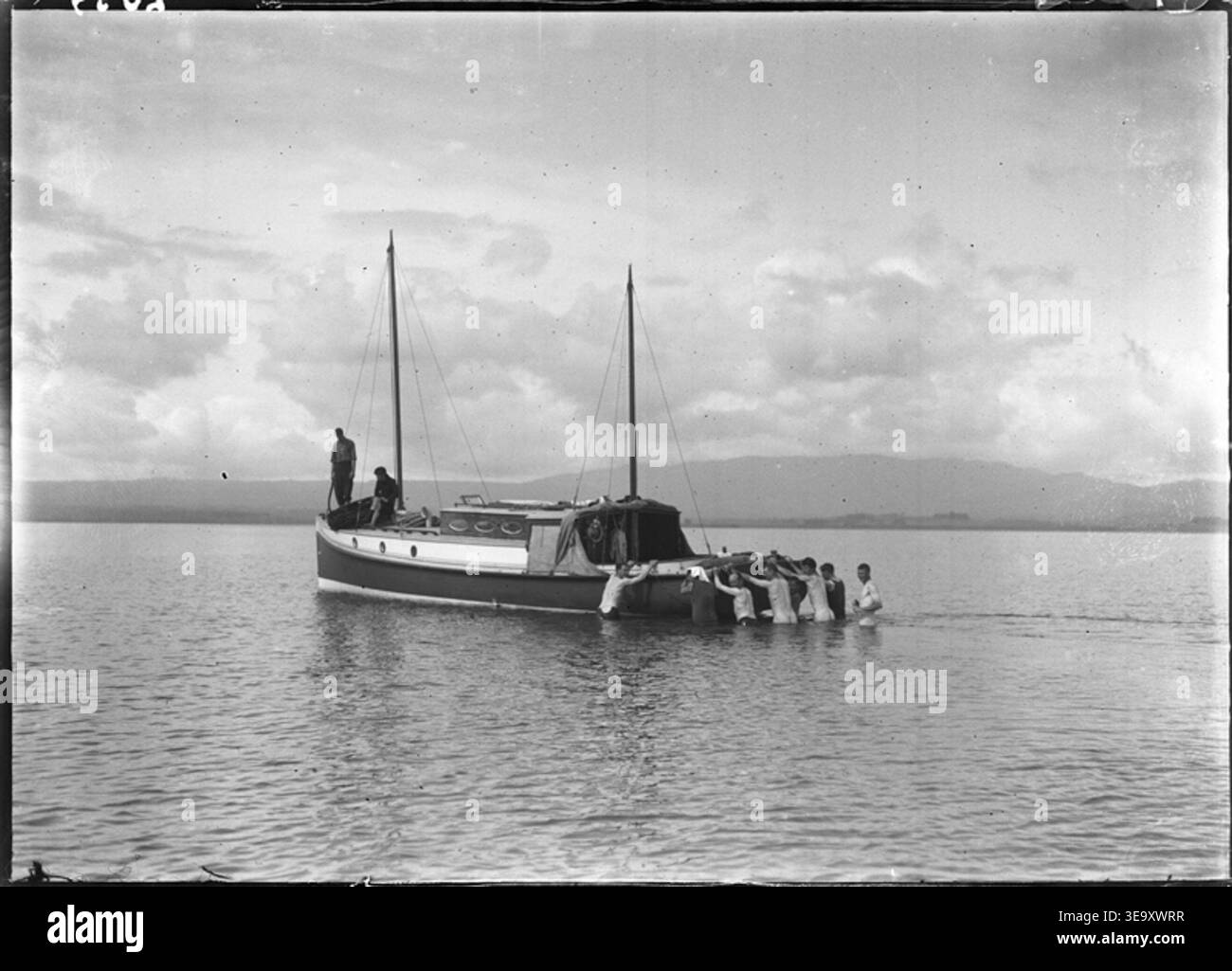 Cette photographie capture le bateau à moteur Uenuku qui s'échoue dans les appartements près de Katikati, en Nouvelle-Zélande, pendant la croisière Matareka. L'image, prise par Henry Winkelmann, montre plusieurs hommes dans l'eau qui tentent de libérer le navire, illustrant les conditions difficiles de la navigation au début du XXe siècle. Banque D'Images