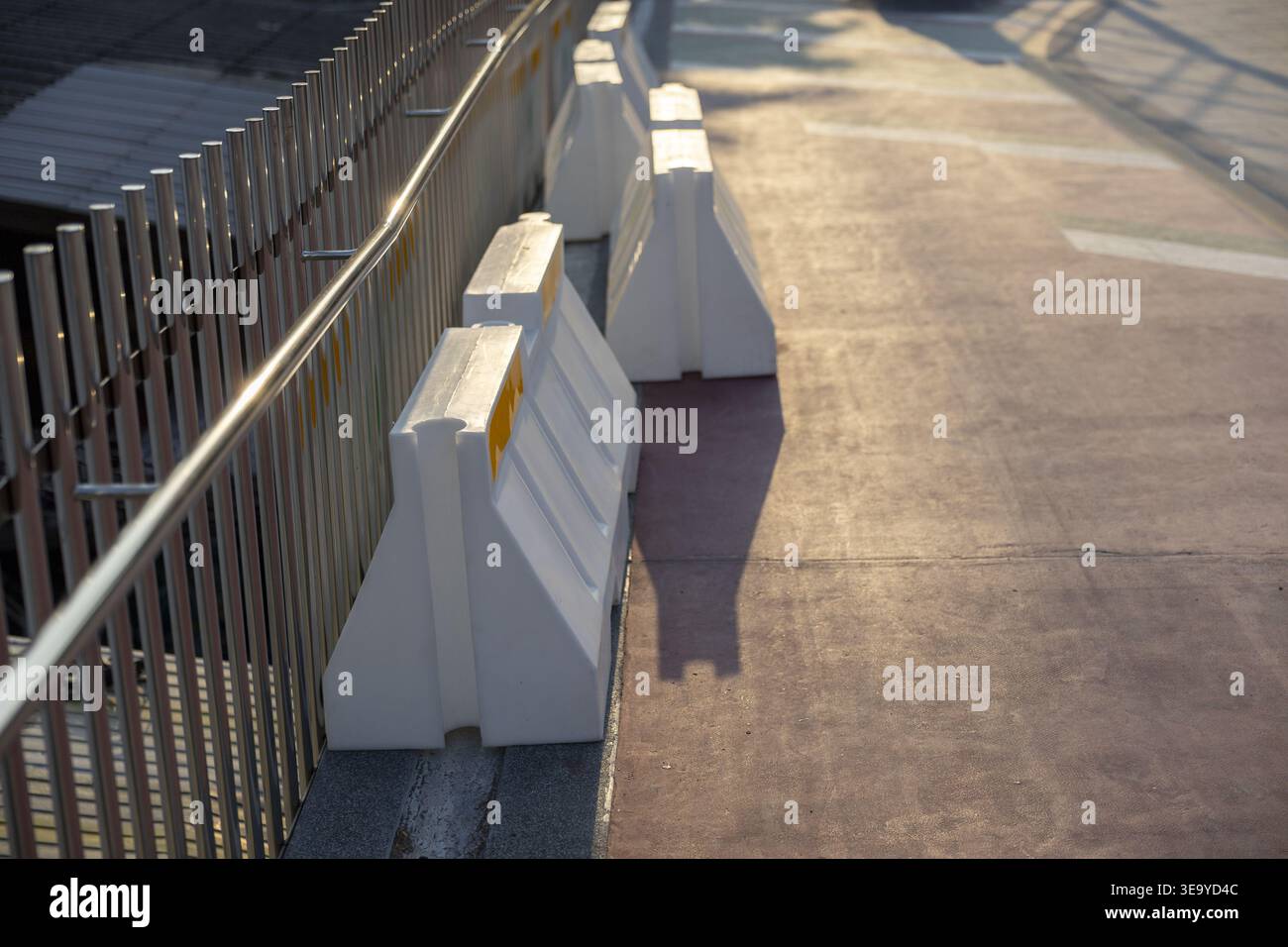 Rangée de barrière de béton en plastique blanc le long de la surface de route asphaltée de clôture métallique pendant le matin ensoleillé brillant coulant de longues ombres offrant un sentiment de sécurité Banque D'Images