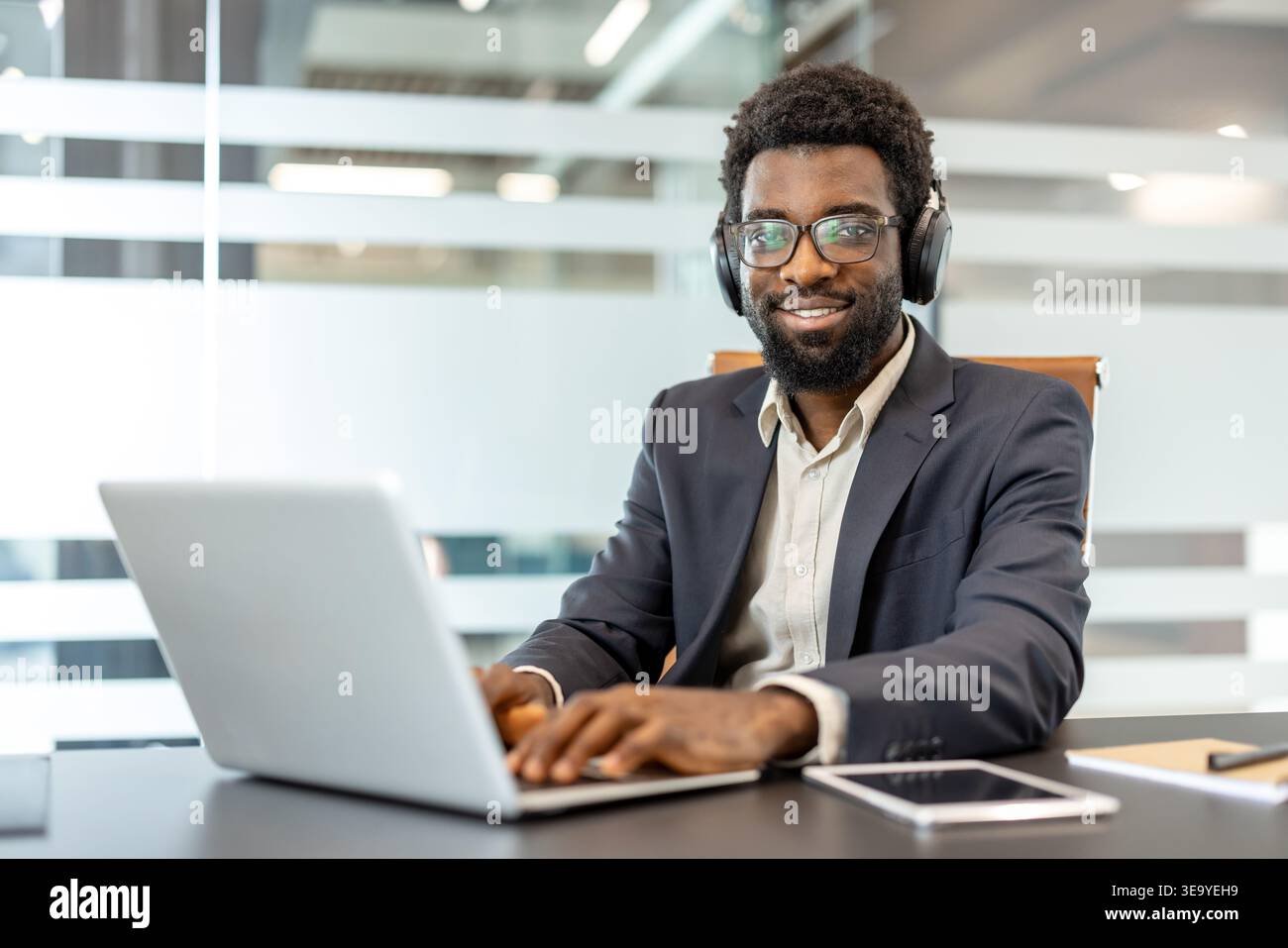 Homme professionnel portant des lunettes et des écouteurs, souriant tout en tapant sur un ordinateur portable à un bureau, symbolisant le travail à distance productif et la technologie de communication moderne Banque D'Images