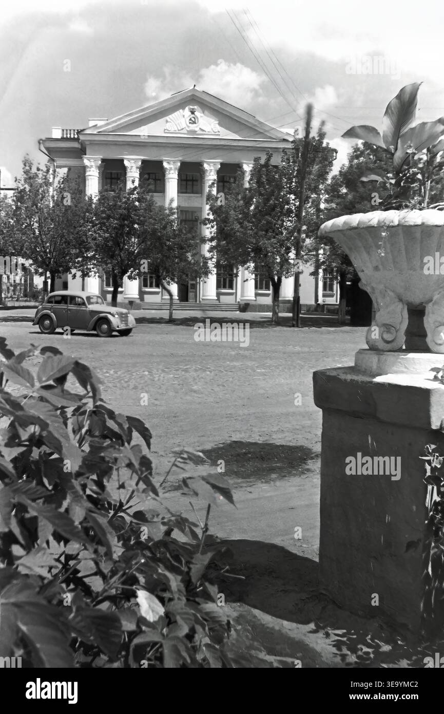 Sloviansk, 1956-1960, rue Svobody. Cette photographie d'archives historiques présente l'architecture classiciste monumentale du collège agricole local. Le grand bâtiment se distingue par un portique massif avec de hautes colonnes cannelées et un emblème soviétique proéminent sur le fronton. Une voiture Moskvitch vintage classique est garée sur la large rue non pavée en face de l'établissement d'enseignement. Les urnes en pierre décoratives et les jeunes arbres plantés ajoutent de l'élégance au paysage urbain du paisible Donbass. Banque D'Images