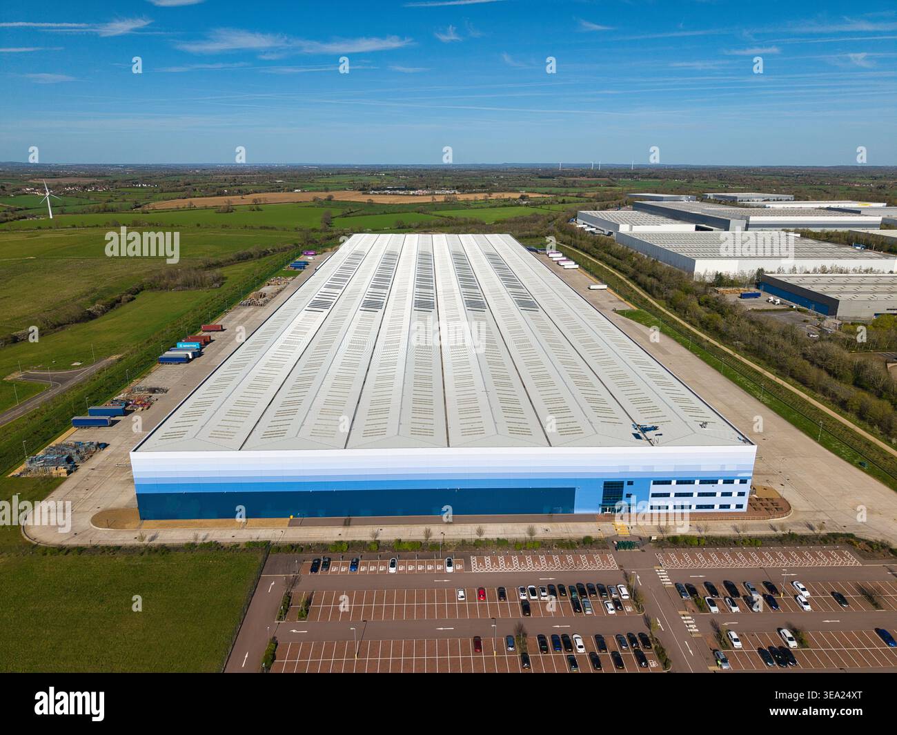 Vue aérienne du grand entrepôt moderne avec façade bleue et vaste aire de stationnement entourée de champs verts Magna Park, Angleterre, United King Banque D'Images