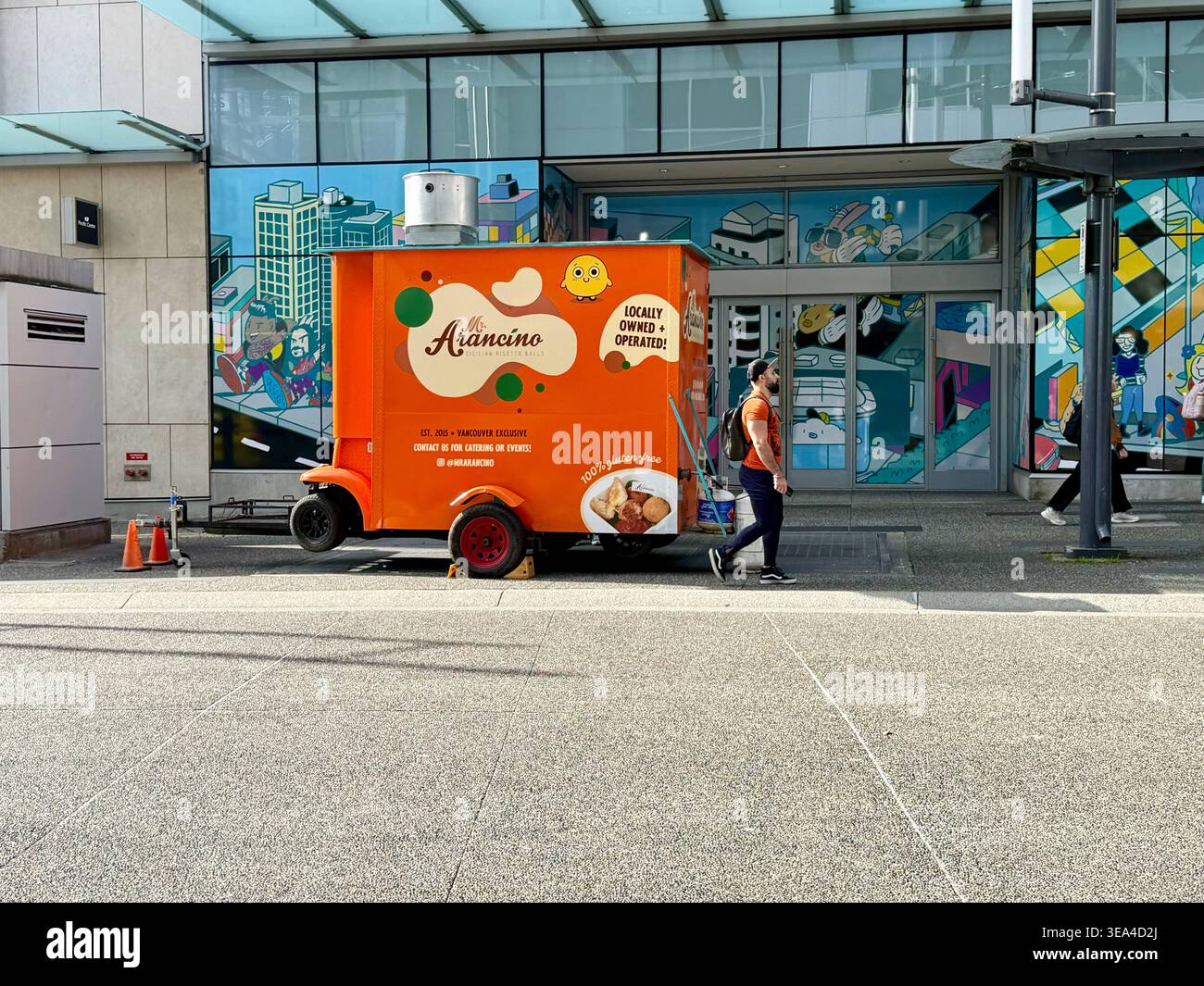 Une scène extérieure vibrante montrant un camion de nourriture « Orange » coloré et un véhicule de livraison garé devant un bâtiment commercial urbain moderne à Vancouver Banque D'Images