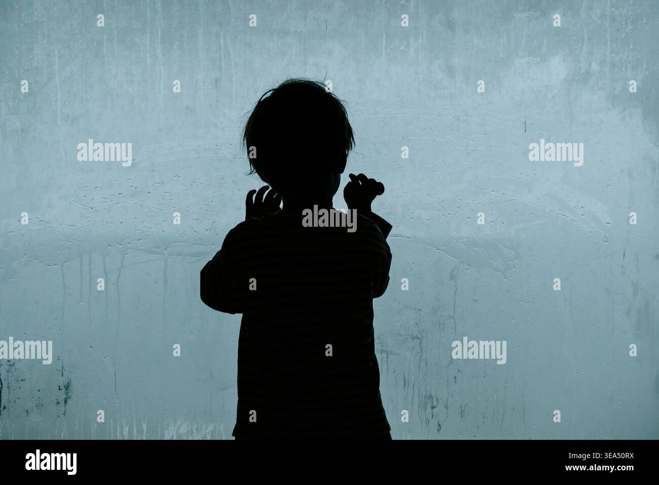 Silhouette d'un enfant solitaire regardant à travers une grande fenêtre embuée froide. Un travail de photographie conceptuelle explorant les thèmes de la dépression infantile, de la crise psychologique et du profond sentiment d’isolement et de tristesse. Photo de haute qualité Banque D'Images