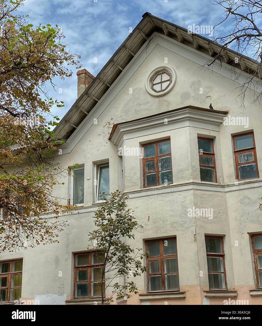 Ancienne façade de bâtiment résidentiel avec fenêtres vintage et arbres d'automne sous ciel nuageux texturé - Image de stock capturée avec un smartphone