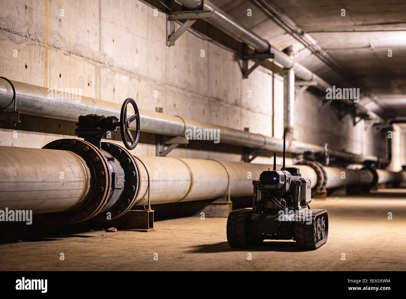 Un petit robot à chenilles avec antennes et caméra est positionné dans un tunnel industriel sombre à côté de tuyaux et de vannes de grand diamètre, suggérant inspec Banque D'Images