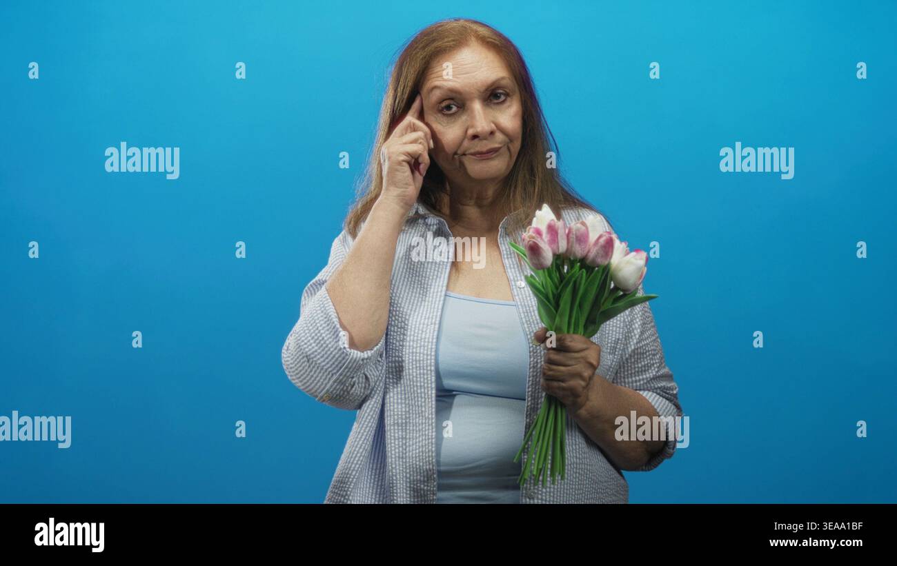 Une femme âgée tenant des tulipes roses touche sa tempe puis pointe le doigt dans le studio ; réflexion réfléchie. Banque D'Images