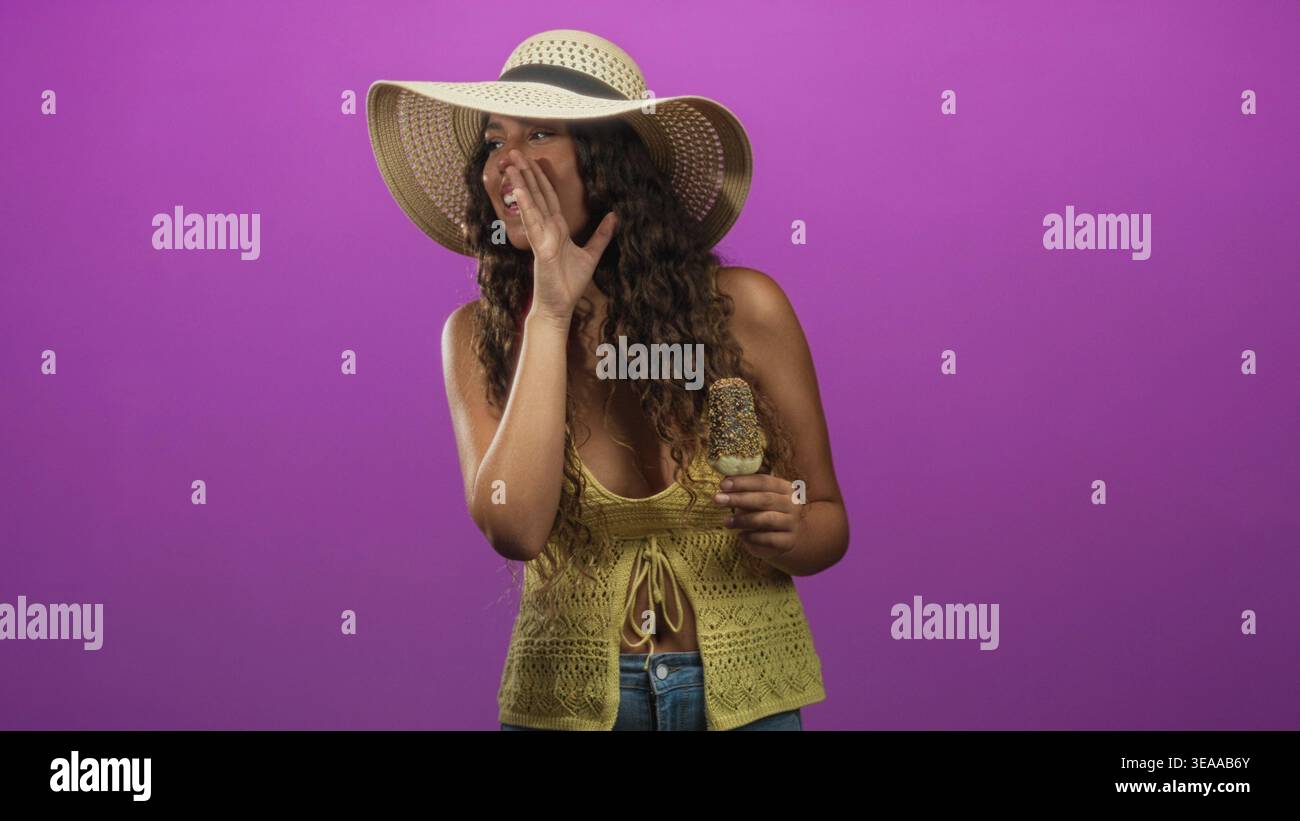 Femme hispanique tient un popsicle saupoudré, main à bouche murmure dans le studio ; vibes d'été secrètes et ludiques. Banque D'Images