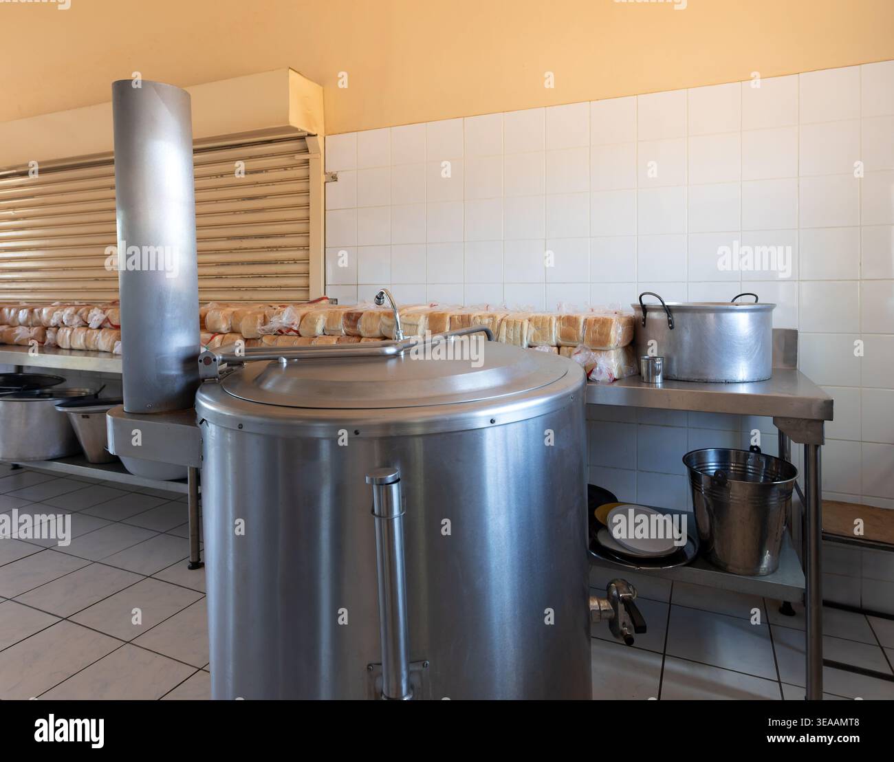 une grande casserole en acier inoxydable à jaquette vapeur se trouve dans une cuisine institutionnelle avec des miches de pain blanc empilées le long d'un comptoir contre un carrelage Banque D'Images