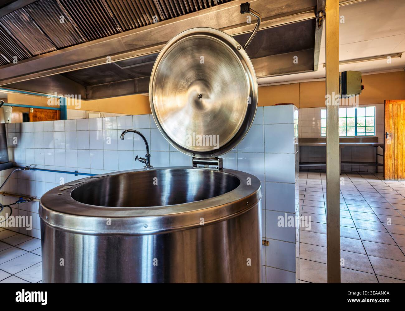 une grande casserole d'ébullition ouverte à chemise de vapeur en acier inoxydable se trouve dans une cuisine institutionnelle carrelée équipée d'une hotte de ventilation robuste et d'un robinet. Banque D'Images
