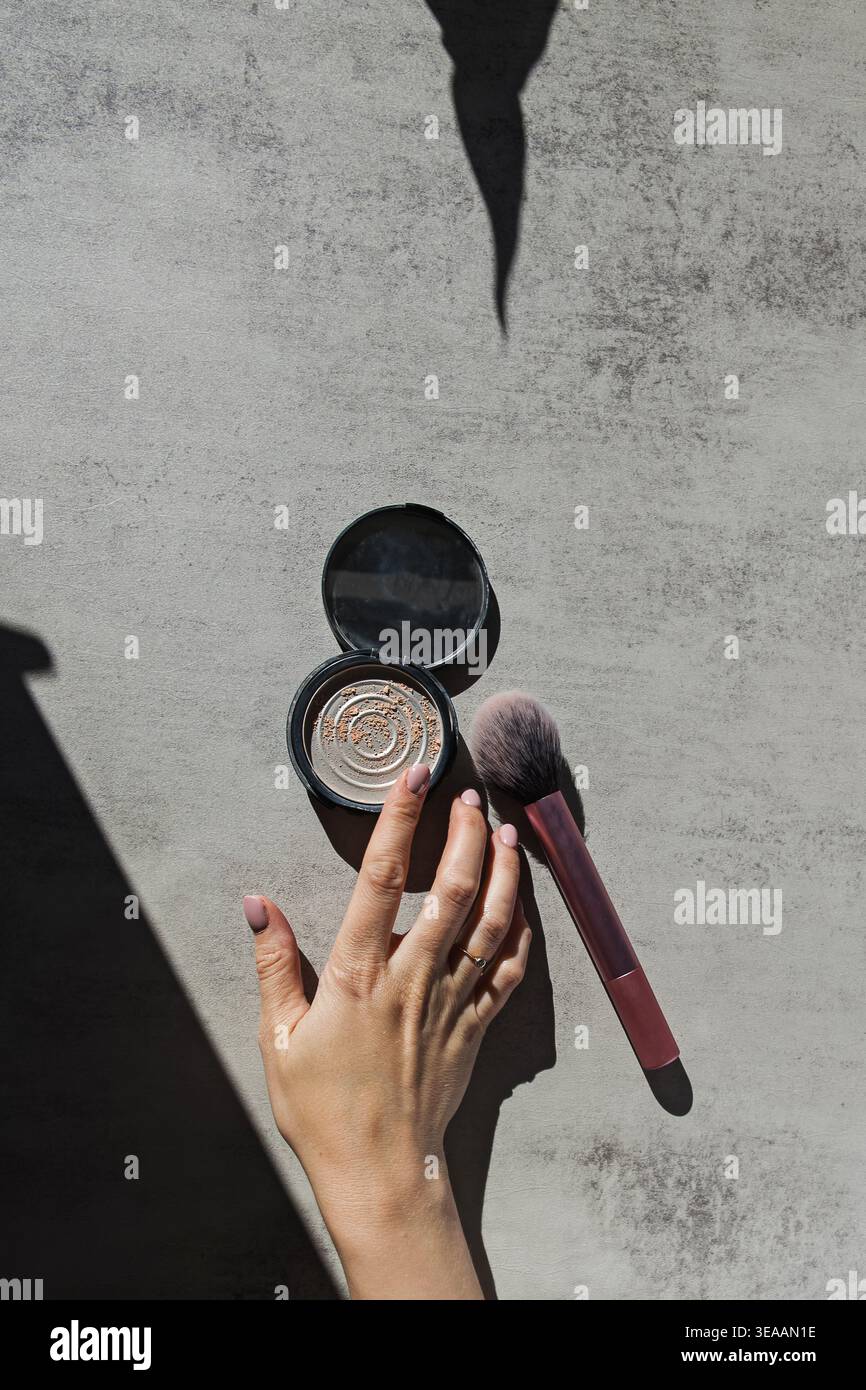 Une main atteint pour un compact presque épuisé poudre à côté d'un pinceau de maquillage sur une surface texturée, minimalisme dans le concept de maquillage Banque D'Images