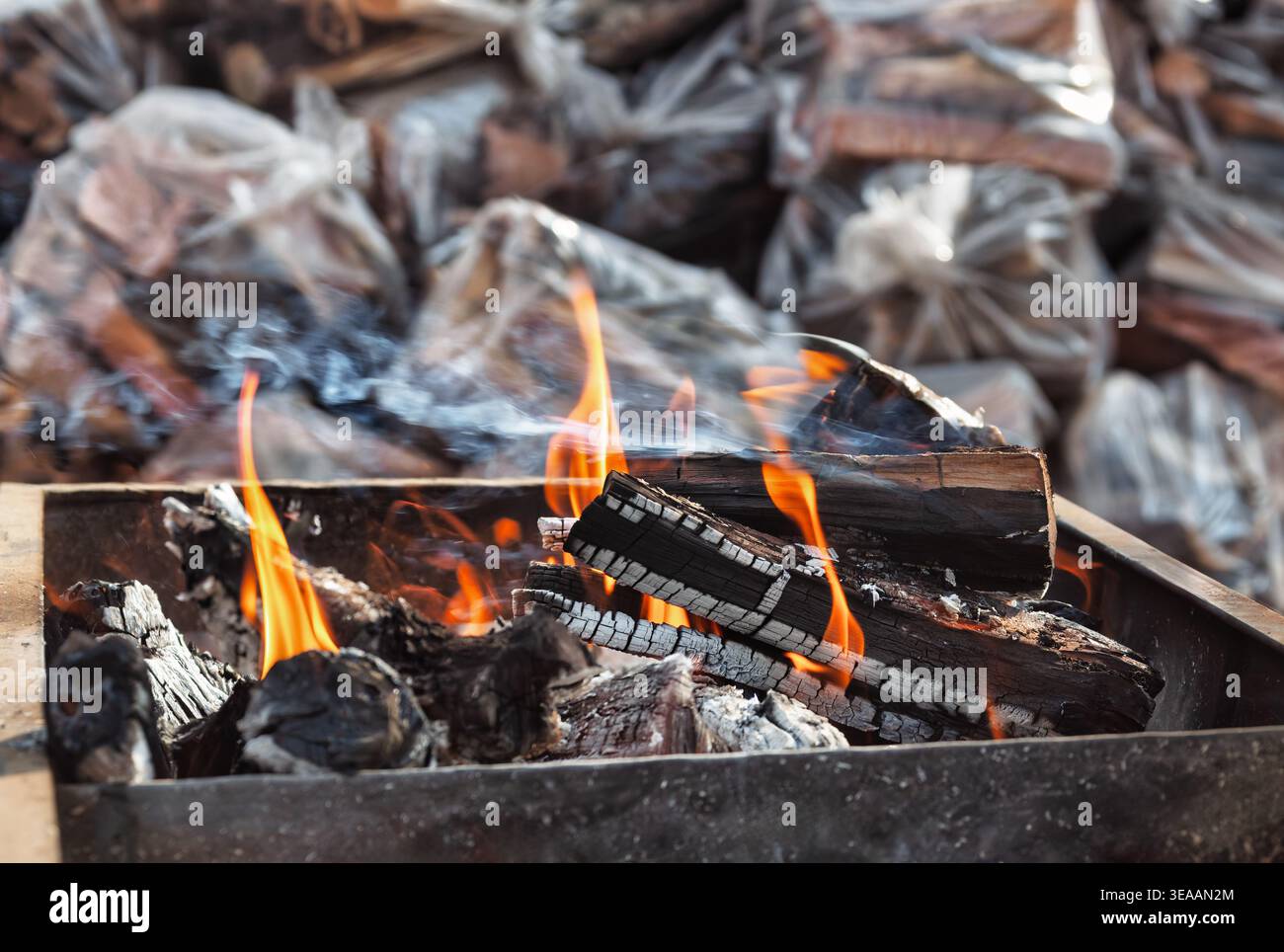 des flammes et de la fumée blanche s'élèvent des bûches carbonisées à l'intérieur d'un foyer en métal foncé avec plusieurs sacs en plastique de bois fendu en arrière-plan. Banque D'Images